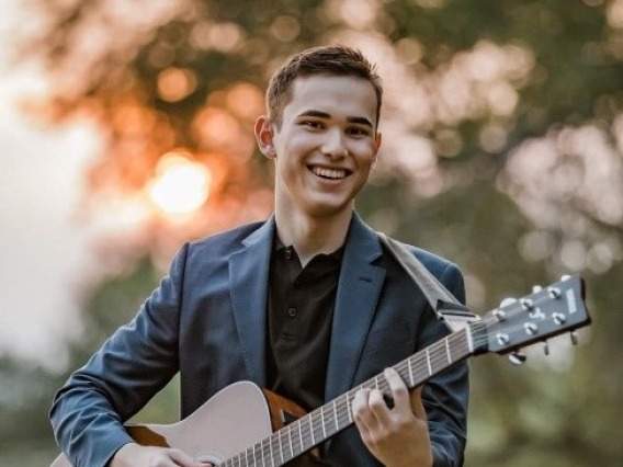 Ayden Carpenter wearing jeans, a black shirt and a sports jacket holding a guitar smiling 