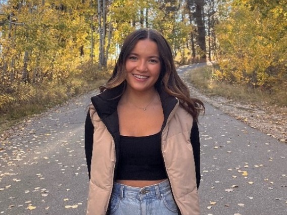 Brianna Hernandez in a tan puffer vest, a black shirt and jeans smiling with trees in the background. 
