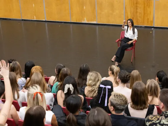 A photo of a woman sitting in a red chair answering students' questions while they sit in a lecture room 