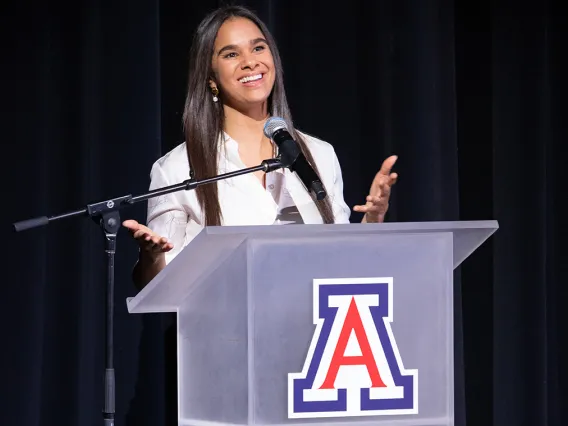 A photograph of a woman looking forward, smiling and talking at a podium 