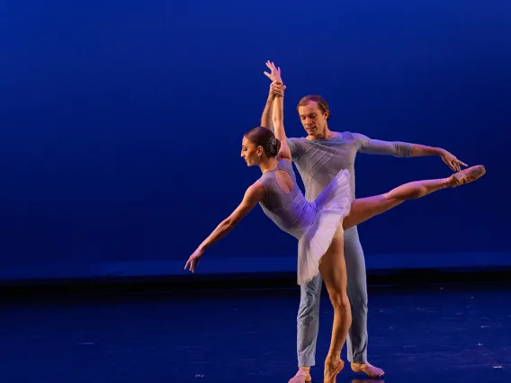 A photograph of one dancer on her tiptoes, being held up by another dancer stretching his body 