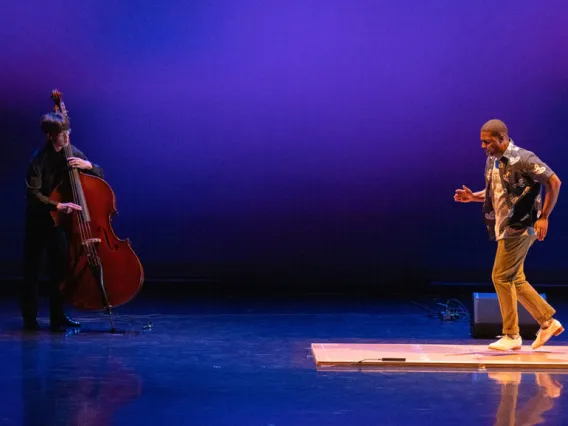 A photograph of a man on the left side plucking away on a cello and a man on the right side tap dancing on a wooden plank on stage 