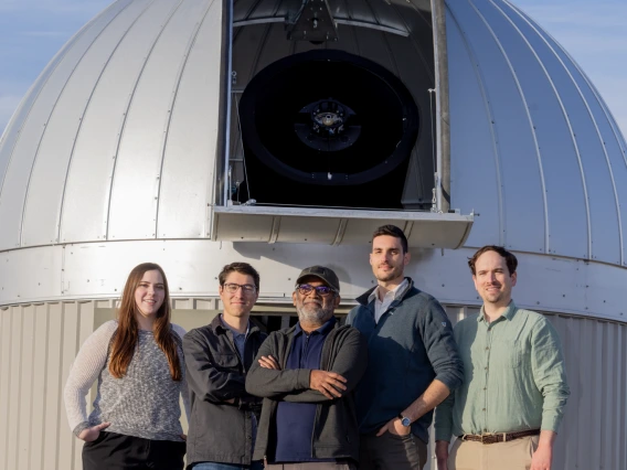 A group of people, one woman and four men, standing and smiling in front of a telescope outside