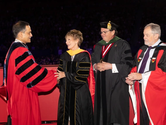 A man in a red academic robe shaking the hand of a woman in a black academic robe, with two other people in academic robes standing behind them. 