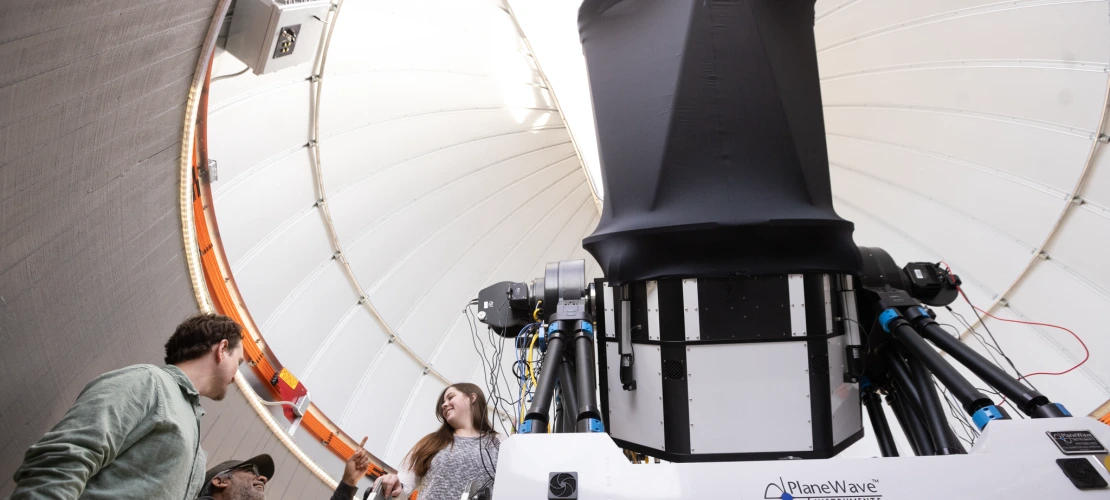 Students and a staff member talking while a student stands on a ladder near a telescope 