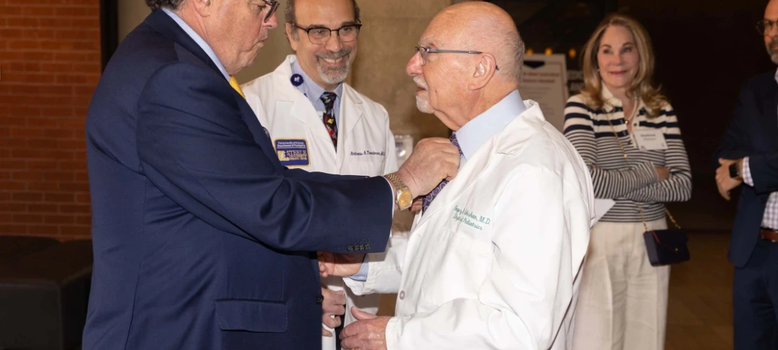 A man adjusting the tie of another, wearing a white coat, while three people smile 
