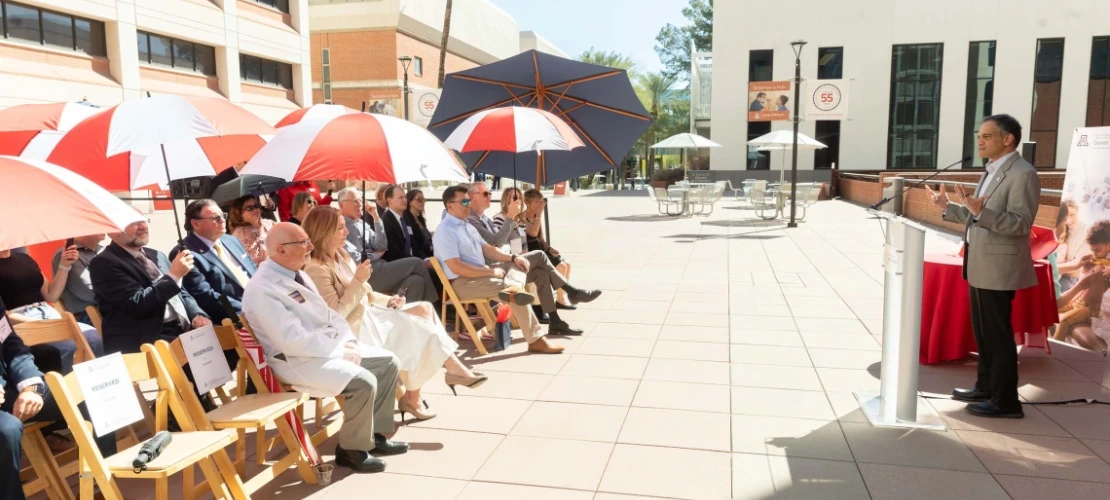 People sitting outside under an umbrella while President Garimella speaks.