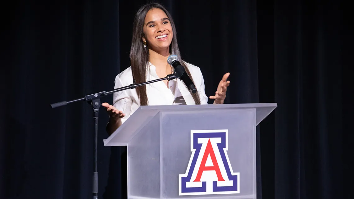 A photograph of a woman looking forward, smiling and talking at a podium 