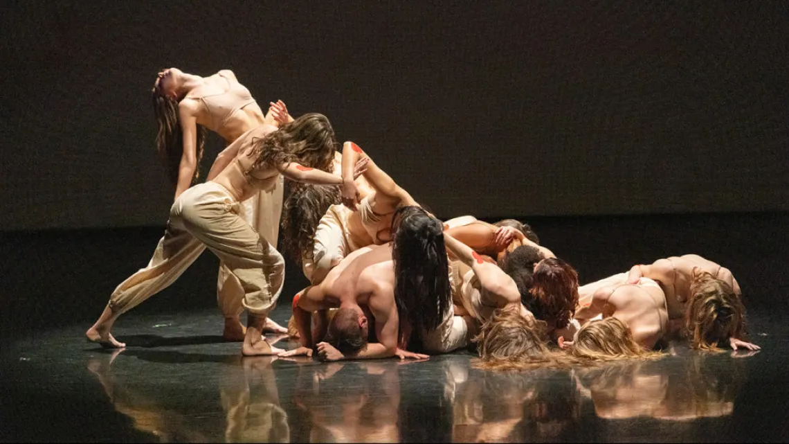 A photograph of dancers wearing tan, draped over each other, on stage 