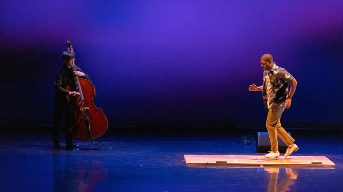 A photograph of a man on the left side plucking away on a cello and a man on the right side tap dancing on a wooden plank on stage 