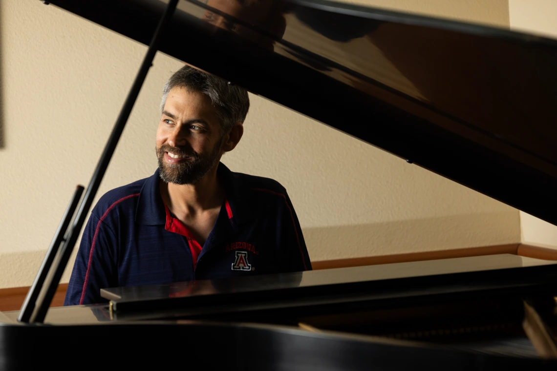 Image of Matthew Mugmon sitting and presumably playing the piano. He is looking to the side and smiling.
