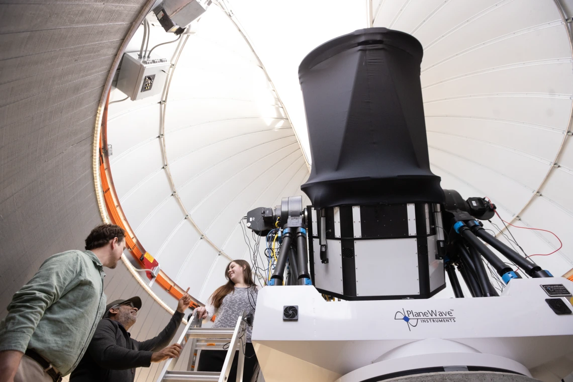 Students and a staff member talking while a student stands on a ladder near a telescope 