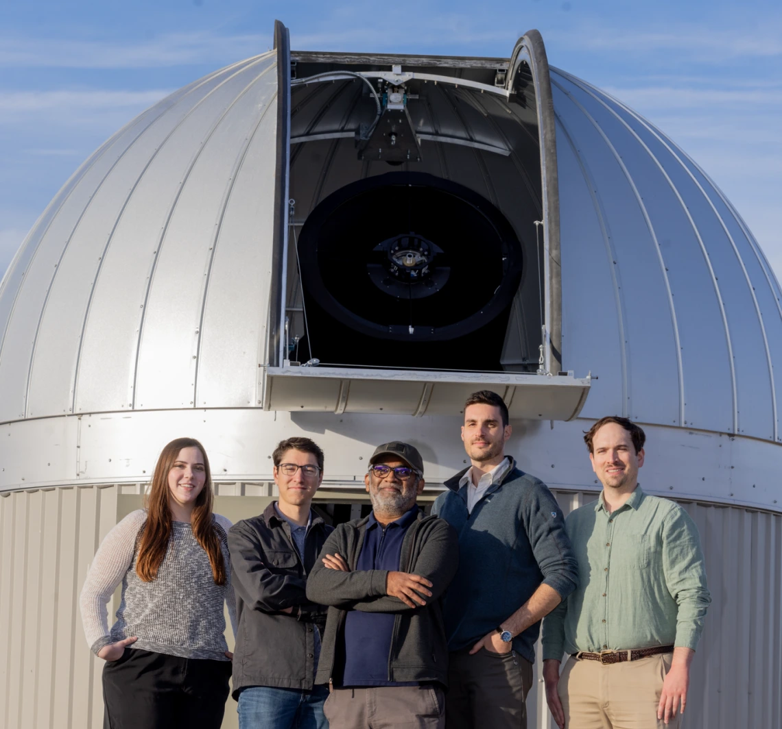 A group of people, one woman and four men, standing and smiling in front of a telescope outside