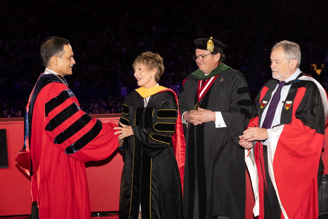 A man in a red academic robe shaking the hand of a woman in a black academic robe, with two other people in academic robes standing behind them.