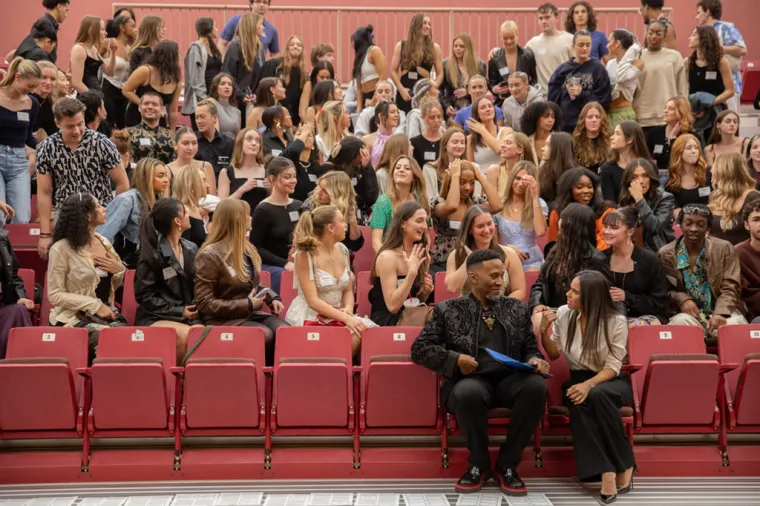 A photograph of a man and woman deep in conversation on red chairs with dozens of students behind them talking, smiling and watching.