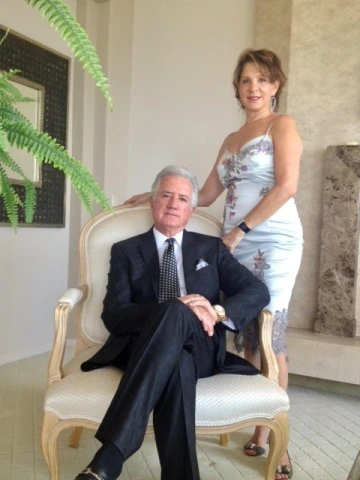 A man sitting with his legs crossed in a chair and a woman standing next to him with her hand on his shoulder.