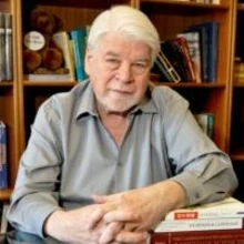 A man smiling with his hands crossed, in front of a book shelf